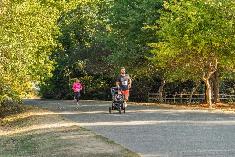Man jogging with stroller in scenic park, woman running behind on a sunny day.
